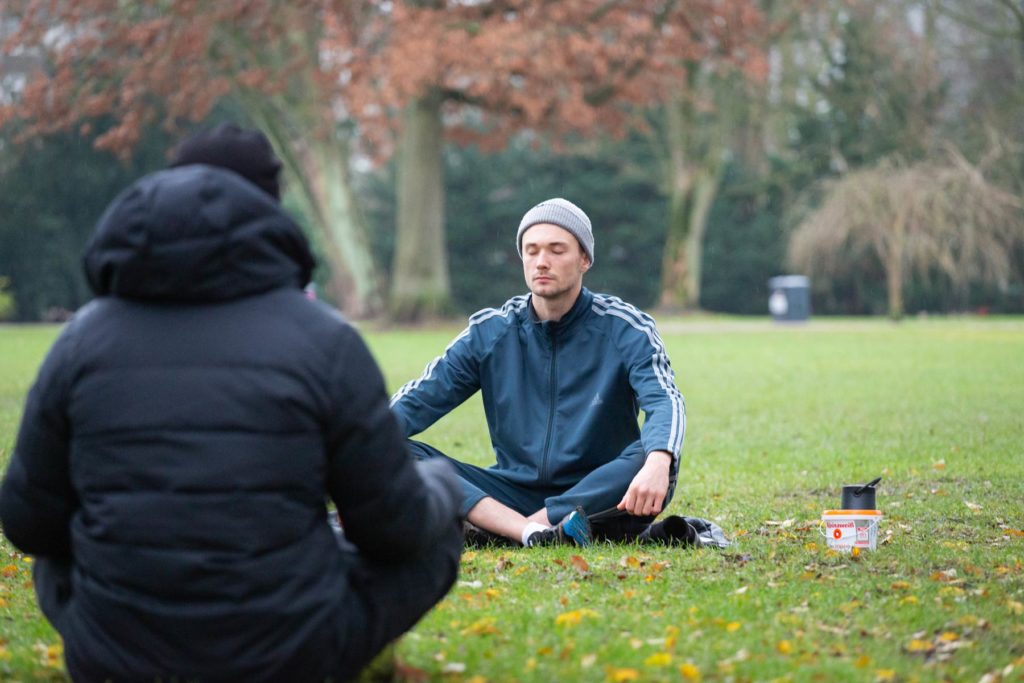 Joshua Alsen und eine Teilnehmende sind dabei zu sehen wie sie im Schneidersitz auf dem Rasen sitzen. Joshua hat seine Augen geschlossen.