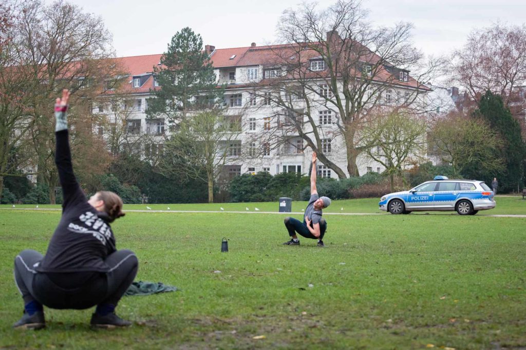 Joshua Alsen und eine Teilnehmende sind auf einer Wiese zu sehen, wie sie in der Hocke einen Arm in die Luft strecken. Im Hintergrund ist ein Streifenwagen der Polizei zu sehen.