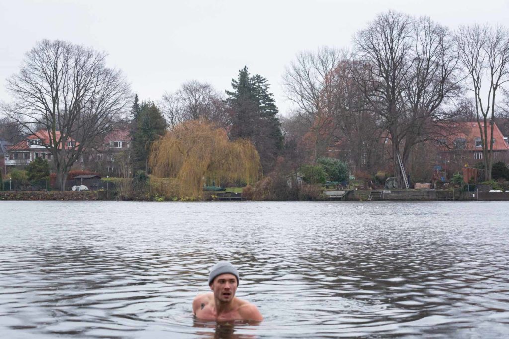 Joshua Alsen ist bis zu den Schultern zu sehen, wie er in der Alster steht. Er trägt eine graue Mütze und blickt zum rechten Bildrand. Im Hintergrund sind Bäume und Häuser zu erkennen.